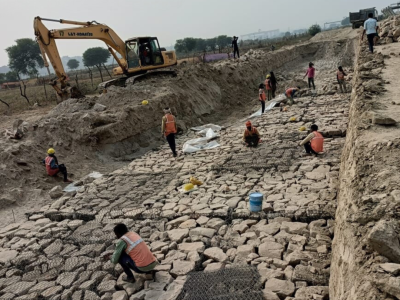 An excavation site with many Indian construction workers laying large stones and mesh on a sloped surface, with an excavator at the top.