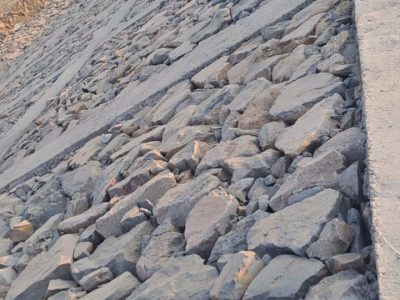 A steep embankment, likely for a dam or canal, is reinforced with a rough arrangement of large, irregularly shaped stones, with a concrete edge and a distant flat landscape.