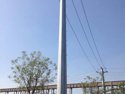 2 A tall, light-colored utility pole with power lines extends into a clear blue sky, with a bridge structure and trees in the background.