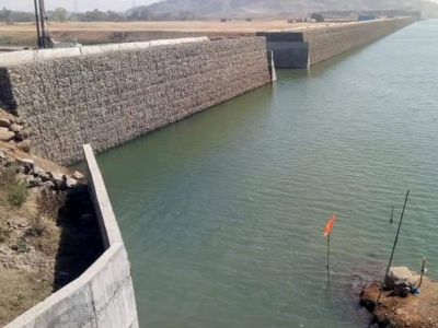 A long, straight canal with gabion walls on one side and a concrete structure on the other, extending towards distant hills under a clear sky.