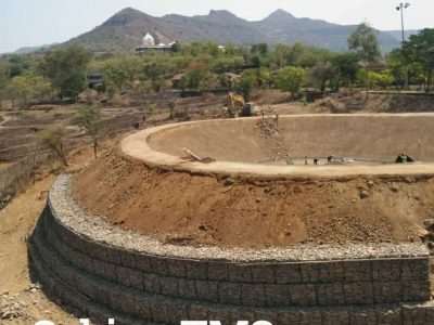 An Indian construction site features a large, excavated circular pit with a tiered gabion wall forming its lower exterior, set against a backdrop of mountains and a temple.