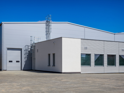 A modern industrial building features a large gray warehouse section with a roll-up door, connected to a smaller white and striped office area, under a bright blue sky.