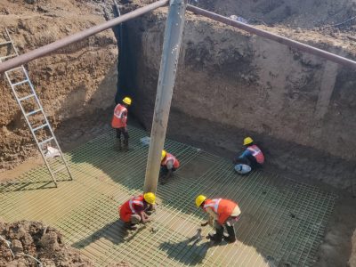 Construction workers in hard hats and safety vests are preparing a concrete foundation with rebar at the bottom of a large trench, with a tractor and pipes nearby.
