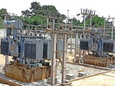 8 Two large grey power transformers with numerous insulators and wires are visible at an outdoor electrical substation, surrounded by gravel and utility structures.