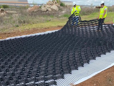 Two construction workers in safety vests and hard hats are unrolling and installing a large sheet of black geocell erosion control matting over a white fabric layer on a sloped, unpaved area.