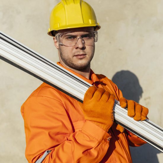 A man in an orange jumpsuit, yellow hard hat, and safety glasses looks at the camera while carrying white pipes or conduits over his shoulder.