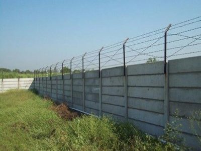 A precast concrete compound wall topped with barbed wire fencing stretches across a grassy area under a clear blue sky.