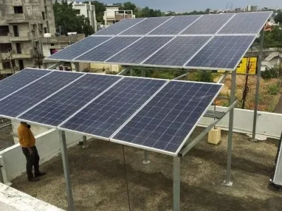 solar-rooftop-power-project-500×500 A residential rooftop in India features two elevated arrays of solar panels, with buildings and trees visible in the background under a cloudy sky.