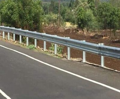 A newly installed galvanized steel crash barrier runs alongside a paved road, with green foliage and trees in the background under a bright sky.